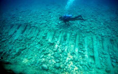 A diver explores underwater ruins with large stone structures and pebbles on the ocean floor.