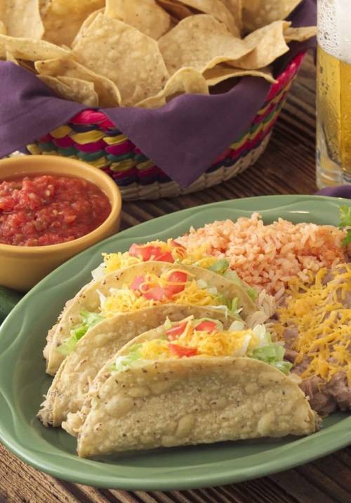 A plate of three tacos with lettuce, cheese, and tomatoes, served with rice, salsa, and a basket of tortilla chips.
