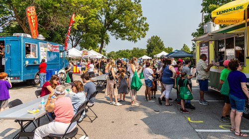 A bustling outdoor food festival with food trucks, tents, and people enjoying food and drinks in a sunny park setting.
