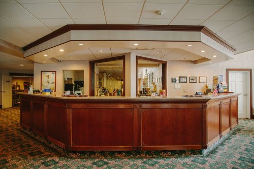 Wooden reception desk in a well-lit lobby with a patterned carpet and various decorations on the walls.
