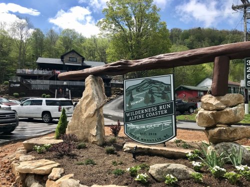 Sign for Wilderness Run Alpine Coaster, surrounded by rocks and greenery, with a building and vehicles in the background.