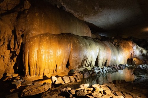 Stalactites and rock formations illuminated in a cave, with a small pool of water reflecting the light.