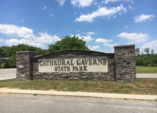 Sign for Cathedral Caverns State Park, featuring stone structure and green landscape under a partly cloudy sky.