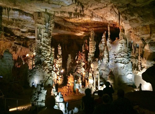 A dimly lit cave interior featuring tall stalagmites and stalactites, with visitors observing the formations.