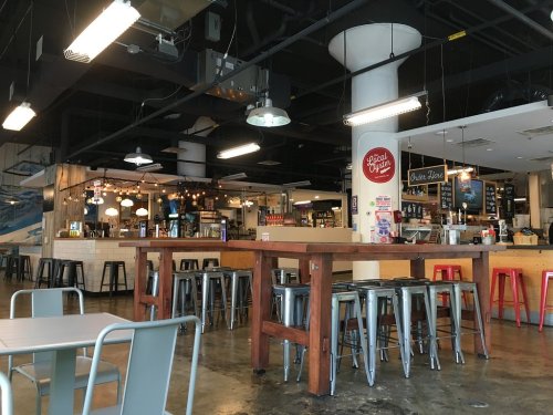 Interior of a modern café with wooden tables, metal stools, and a bar area in the background. Bright and inviting atmosphere.