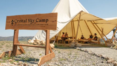A wooden sign for Crystal Sky Camp in front of a large tent with people seated around a table in a scenic outdoor setting.