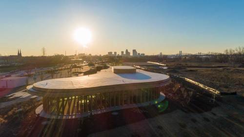 Aerial view of a modern building with a curved roof, set against a sunset and a city skyline in the background.