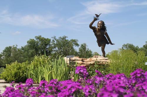 A bronze statue of a girl joyfully reaching for a butterfly, surrounded by vibrant purple flowers and greenery.