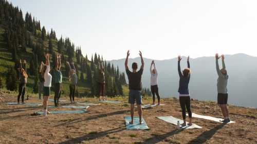 A group of people practicing yoga outdoors on a mountain, stretching with arms raised towards the sky.