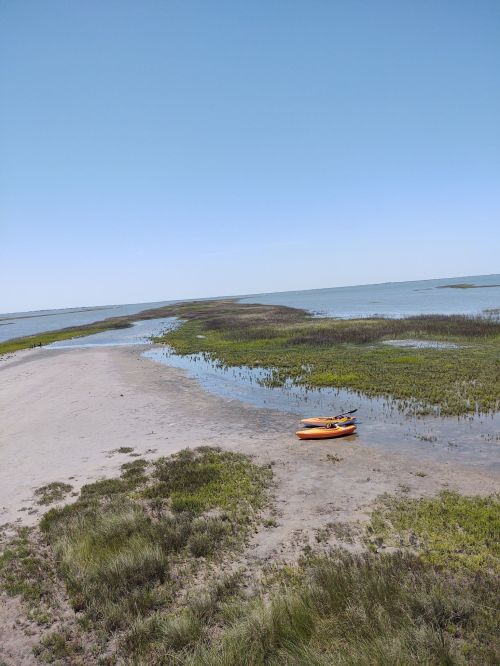 A kayak rests on a sandy shore surrounded by marshland and calm waters under a clear blue sky.