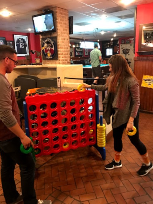 Two people playing a large Connect Four game in a casual bar setting, with a TV and other activities in the background.