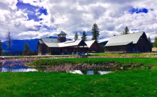 A scenic view of a rustic building by a pond, surrounded by green grass and mountains under a cloudy sky.