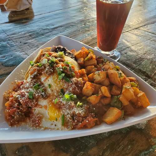 A plate of eggs topped with sauce, cheese, and herbs, served with seasoned potatoes and a glass of drink.