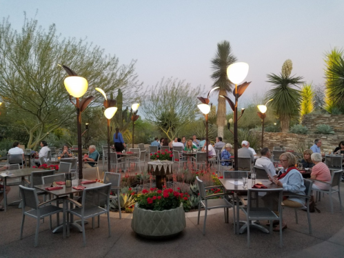 Outdoor dining area with tables, surrounded by plants and decorative lights, during twilight. Guests are enjoying their meals.