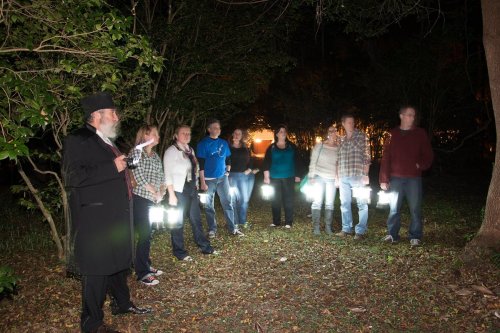 A group of people holding lanterns stands in a dark forest, with one person in a long coat speaking to them.
