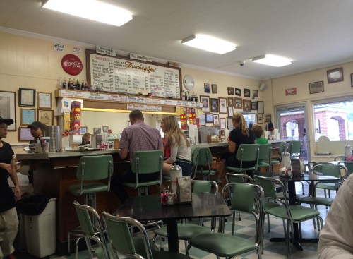 A retro diner interior with patrons at the counter, vintage decor, and a menu board on the wall.