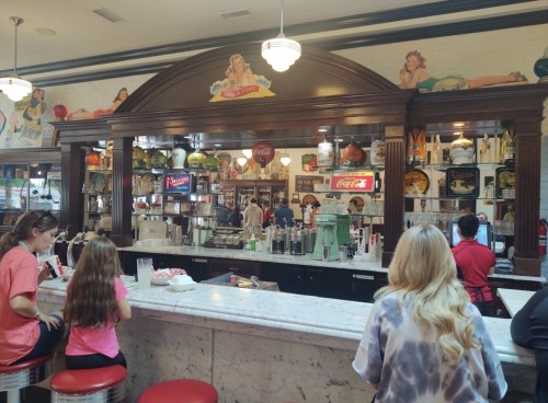 A vintage diner interior with a marble counter, retro decor, and patrons enjoying food and drinks.