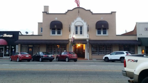 A two-story building with an American flag, featuring arched windows and awnings, surrounded by parked cars.