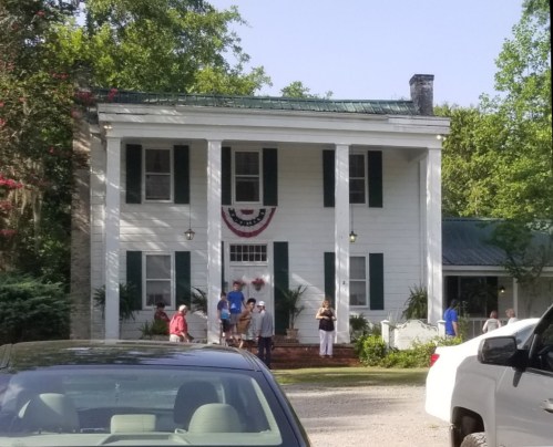 A white two-story house with green shutters, decorated with bunting, and people gathered outside. Cars are parked in front.