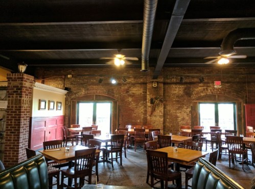 Interior of a rustic restaurant with wooden tables and chairs, exposed brick walls, and ceiling fans.