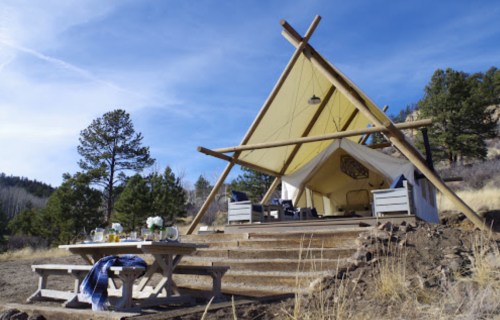 A cozy glamping tent with a wooden deck, surrounded by trees and mountains, featuring a table set for breakfast.