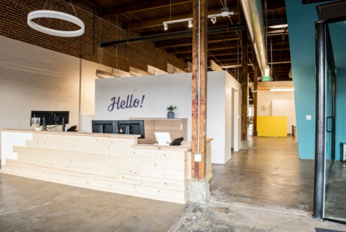 Modern office reception area with a wooden desk, "Hello!" sign, and open layout featuring exposed brick and colorful walls.