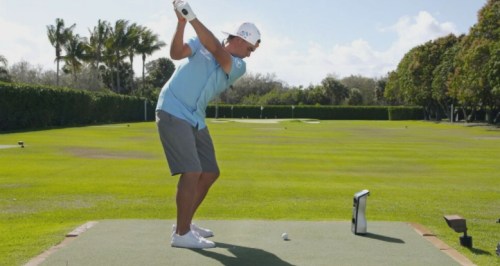 A golfer prepares to swing on a sunny driving range, with palm trees in the background and a golf ball on the tee.