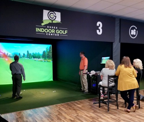 A group of people watches as a man plays golf in a virtual simulator at Essex Indoor Golf Center.