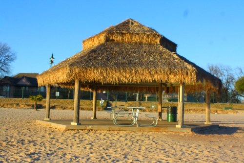 A thatched-roof gazebo on a sandy beach, with a picnic table underneath and trees in the background.