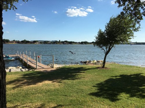 A serene lakeside view featuring a wooden dock, green grass, and a clear blue sky with a few clouds.