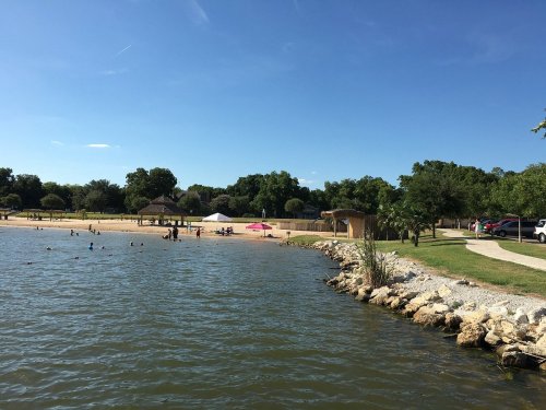 A sunny lakeside scene with a sandy beach, people swimming, and trees lining the shore under a clear blue sky.