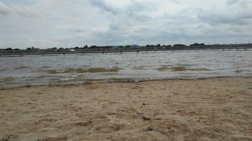 A sandy beach with gentle waves lapping at the shore under a cloudy sky. A pier is visible in the distance.