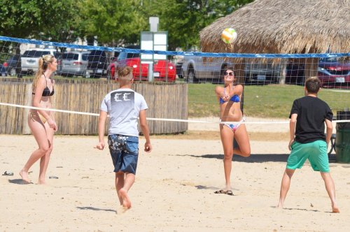Group of four people playing beach volleyball on a sunny day, with a thatched hut and cars in the background.