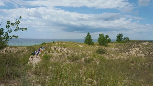 A sandy path leads through grassy dunes to a serene lake under a partly cloudy sky.