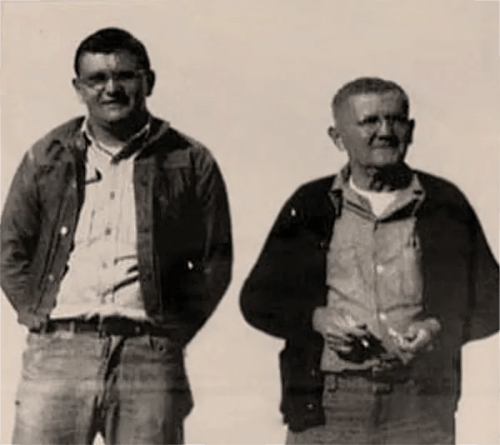 Two men stand side by side, both wearing casual clothing, with a neutral background. One holds an object in his hands.
