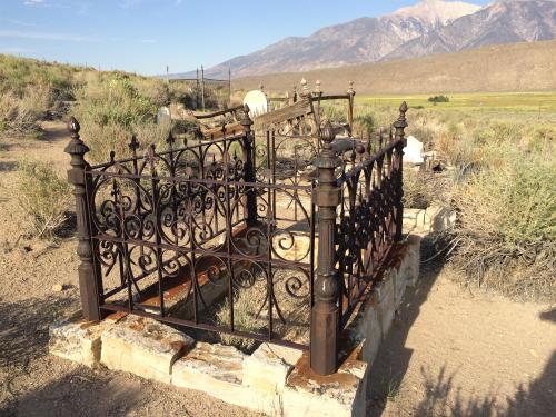 An ornate iron fence surrounds a weathered grave in a desert landscape with mountains in the background.