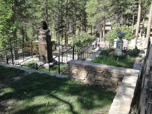 A scenic view of a cemetery with a statue, surrounded by trees and a stone wall, featuring graves and flowers.
