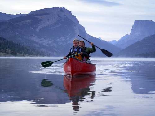 Two people paddle a red canoe on a calm lake, surrounded by mountains and a misty morning sky.