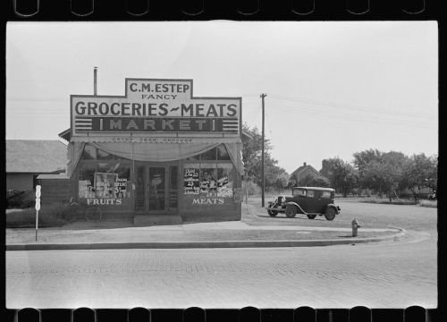 Historic grocery store with a sign reading "C.M. Estep Fancy Groceries-Meats," featuring a vintage car parked outside.