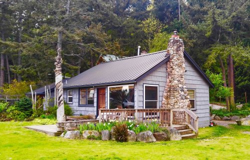 A cozy gray cabin with a stone chimney, surrounded by greenery and a wooden deck in a tranquil setting.
