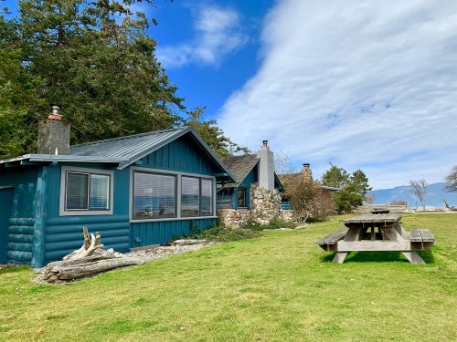A blue cabin with a stone chimney beside a picnic table on a grassy area, under a partly cloudy sky.