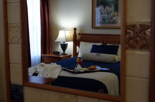 A cozy hotel room with a bed, lamp, and a breakfast tray featuring fruit and pastries, reflected in a mirror.