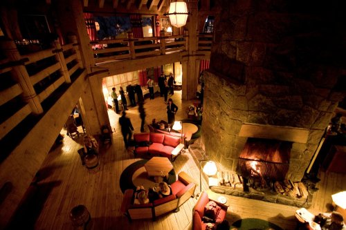 Cozy lodge interior with a large stone fireplace, seating areas, and people socializing in a warm, inviting atmosphere.