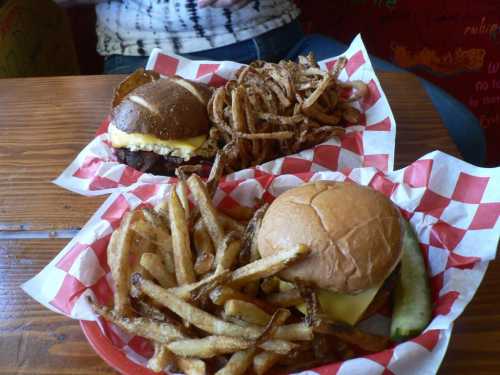 Two plates of food: one with a cheeseburger and crispy fries, the other with a burger and onion rings, all in checkered baskets.