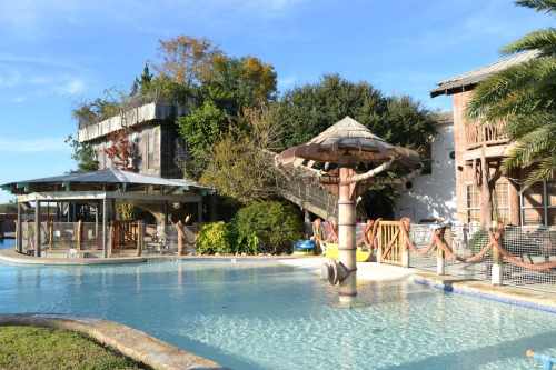 A tropical-themed pool area with a thatched-roof structure, surrounded by lush greenery and wooden buildings.
