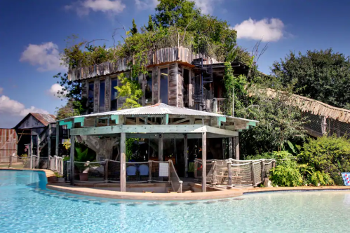 A unique treehouse structure surrounded by lush greenery and a pool, under a bright blue sky.