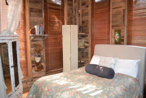 Cozy bedroom with a floral bedspread, wooden walls, and natural light from blinds. Decor includes a mirror and bookshelf.