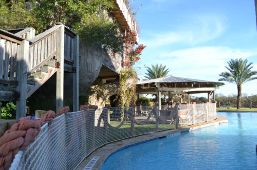 A serene pool area with wooden structures, palm trees, and a shaded seating area under a clear blue sky.