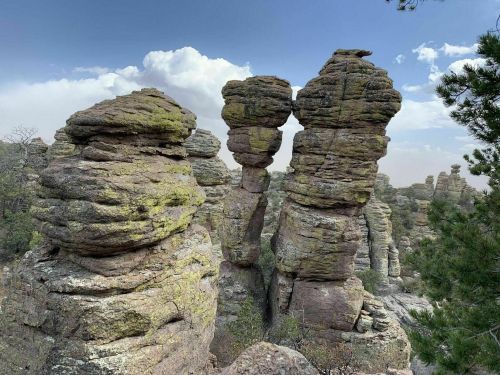 Unique rock formations rise against a blue sky with clouds, surrounded by trees in a natural landscape.