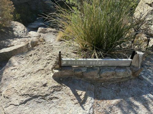 Sign for "Kissing Rocks" on rocky terrain, surrounded by sparse vegetation and a clear blue sky.